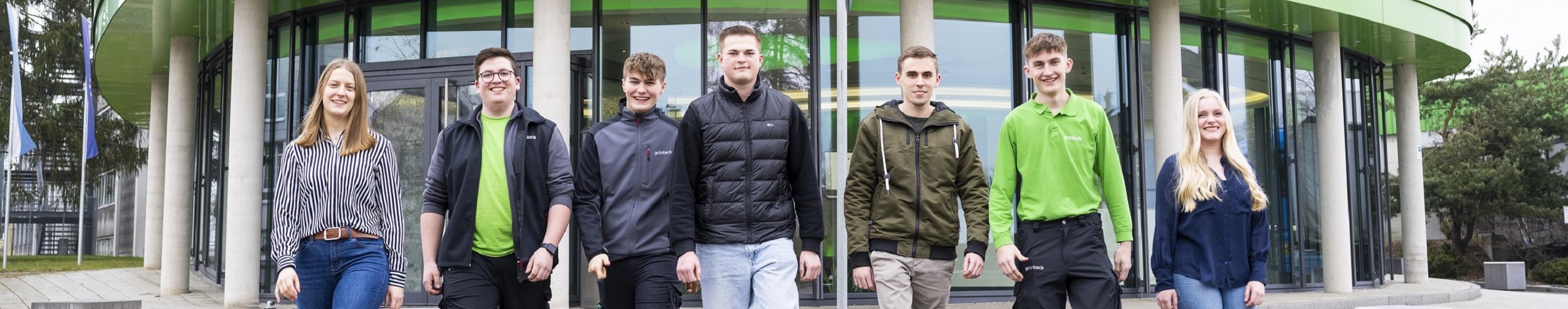 Mehrere Menschen gehen nebeneinander vor einem modernen Gebäude mit Glasfassade und grünen Akzenten.