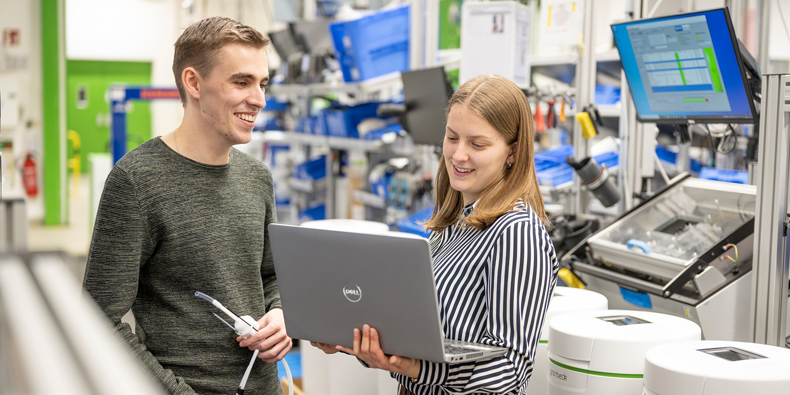 Personen interagieren lächelnd mit einem Laptop in einer technisch ausgestatteten Umgebung mit Monitoren und Geräten im Hintergrund.