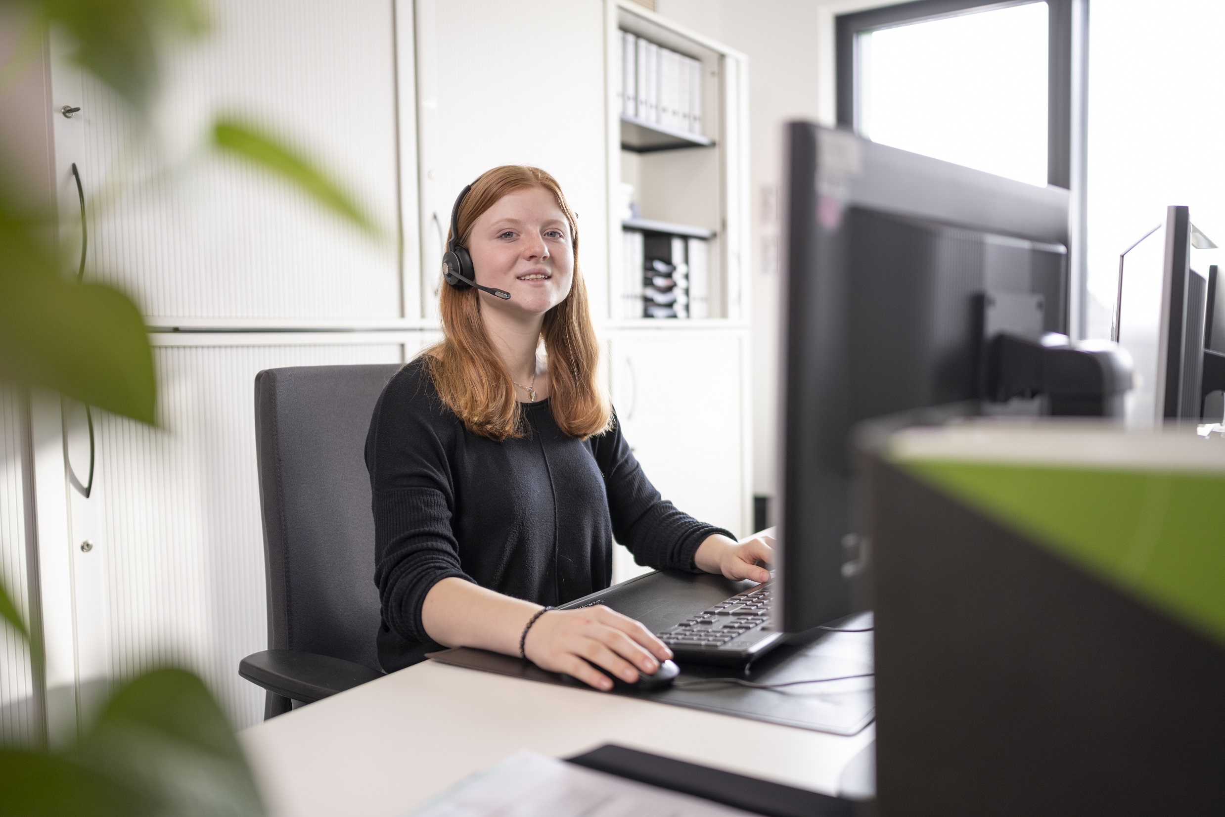 Eine Frau mit Headset sitzt lächelnd vor einem Computer in einem Büro, umgeben von Aktenschränken und Ordnern.